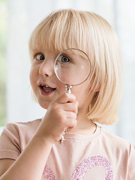 Little girl looking through a magnifying glass with one eye, smiling playfully.